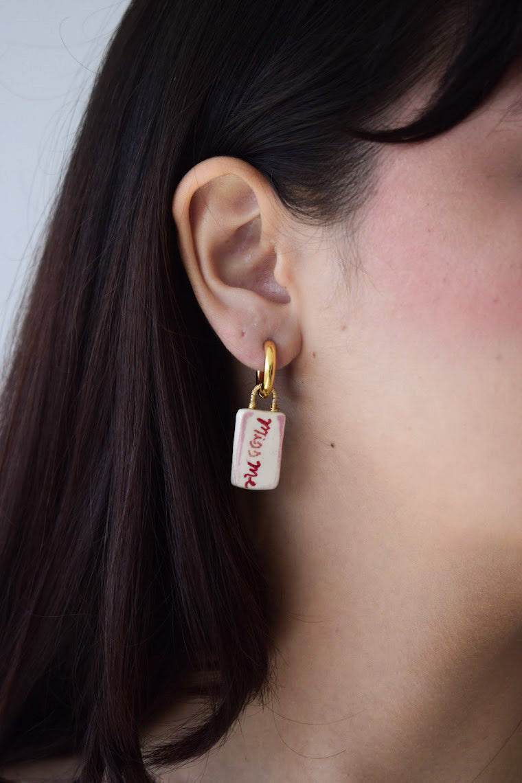 Oreja de modelo de tes blanca y cabello negro usando hoops o candongas “miss me” con dije en cerámica en colores rojo y blanco, mensaje pintado a mano.