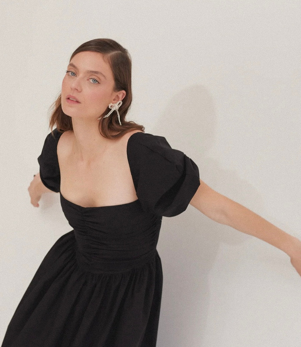 Modelo de tes blanca y cabello castaño en fondo blanco de Sixxta con vestido negro y aretes Teddy’s en color transparente.