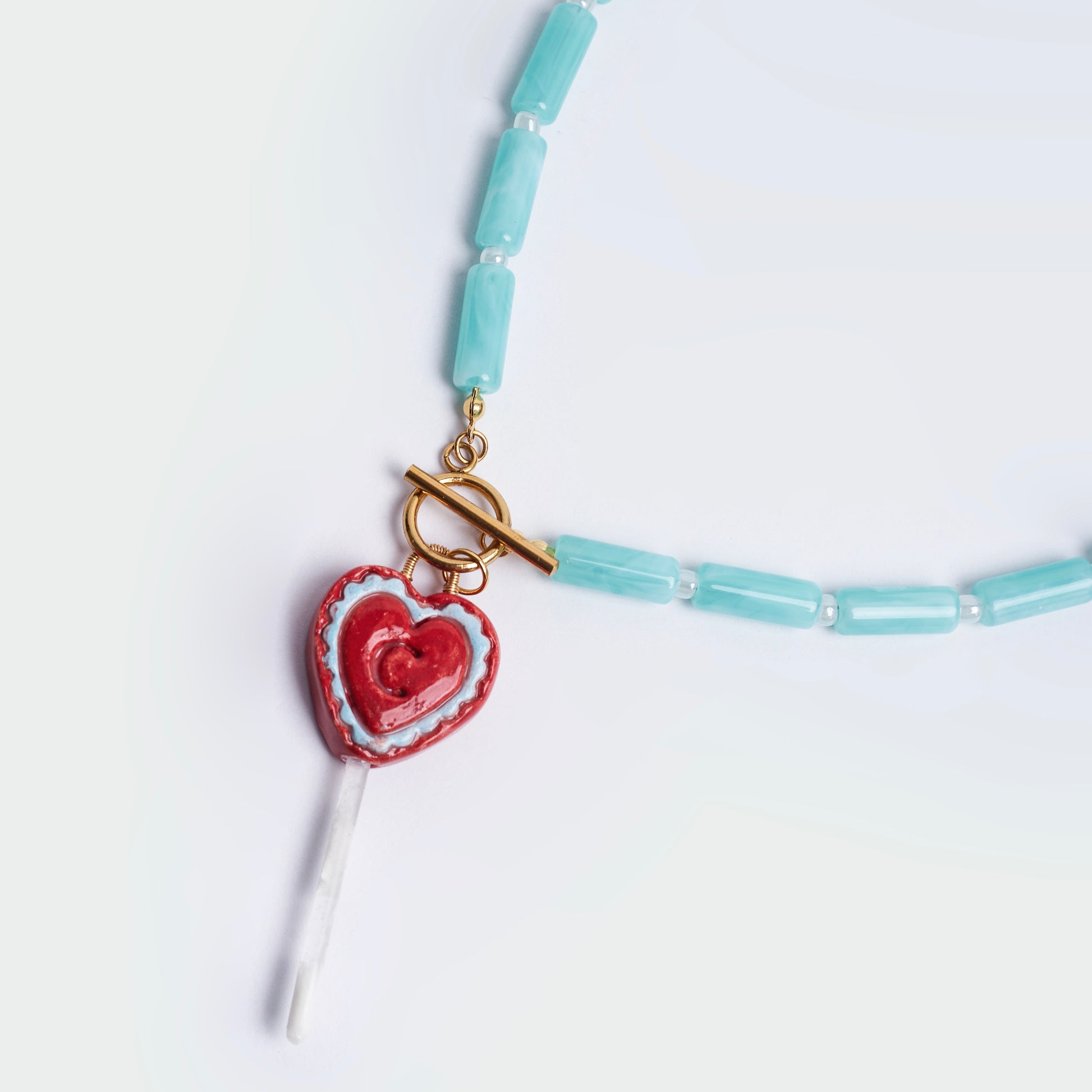 Collar hecho a mano con cuencas cilíndricas color esmeralda, perlas blancas y broche salvavidas en acero inoxidable color dorado, con un dije en forma de chupeta en material de cerámica en colores rojo y celeste y una vara blanca en resina ubicada al final. 