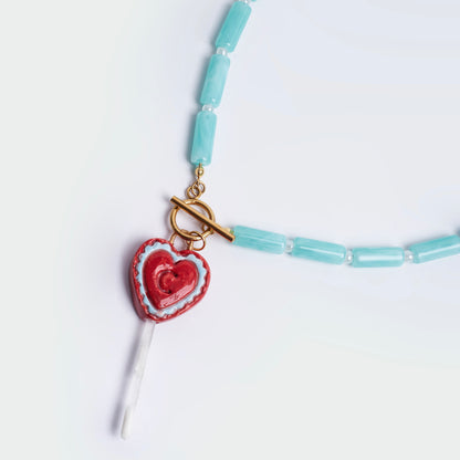 Collar hecho a mano con cuencas cilíndricas color esmeralda, perlas blancas y broche salvavidas en acero inoxidable color dorado, con un dije en forma de chupeta en material de cerámica en colores rojo y celeste y una vara blanca en resina ubicada al final. 