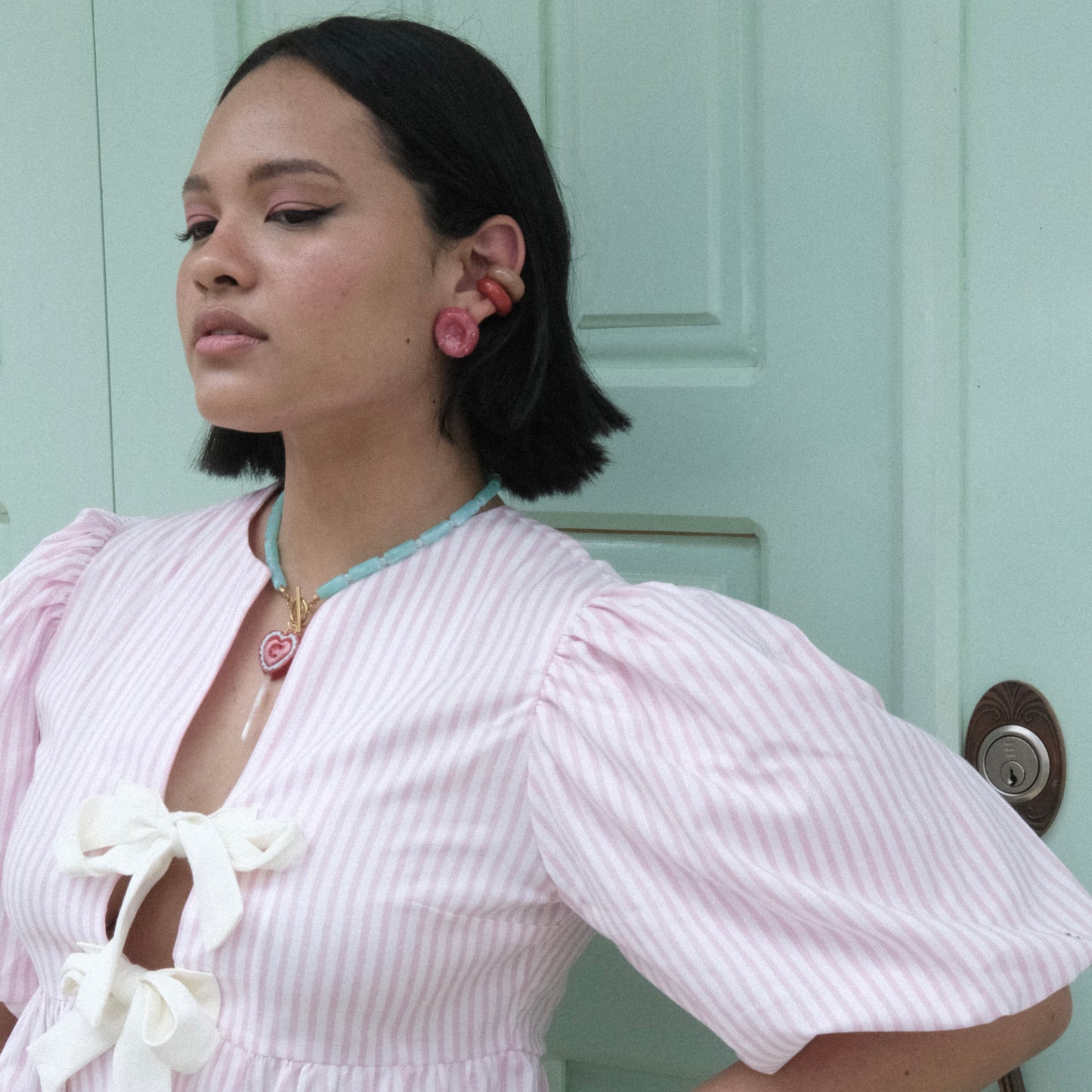 Modelo pluszize de tes morena y cabello corto color negro en fondo de puerta color verde menta, usando top de nudos con rayas rosadas y blancas y set Bombón de Chouette con earcufs en colores rojo, marrón, celeste y verde