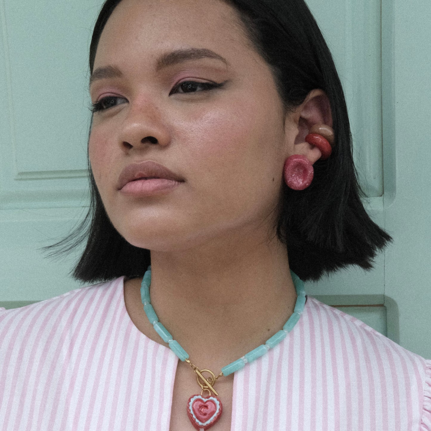 Rostro y torso de modelo pluszize de tes morena, cabello corto y negro en fondo de puerta verde menta, usando top de rayas rosas y blancas, collar Chupipop, aretes Deliciosos y Earcuffs festival clásicos tamarindo y bocadillo. Accesorios en material de cerámica en clores rojo, marrón y celeste