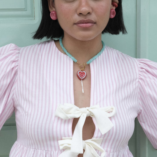 Rostro y torso de modelo pluszize de tes morena, cabello negro y corto usando top de moños coquette en rayas blancas y rosas, collar Chupipop, aretes Deliciosos y earcuffs bombón clásicos en colores rojo, marrón y celeste.