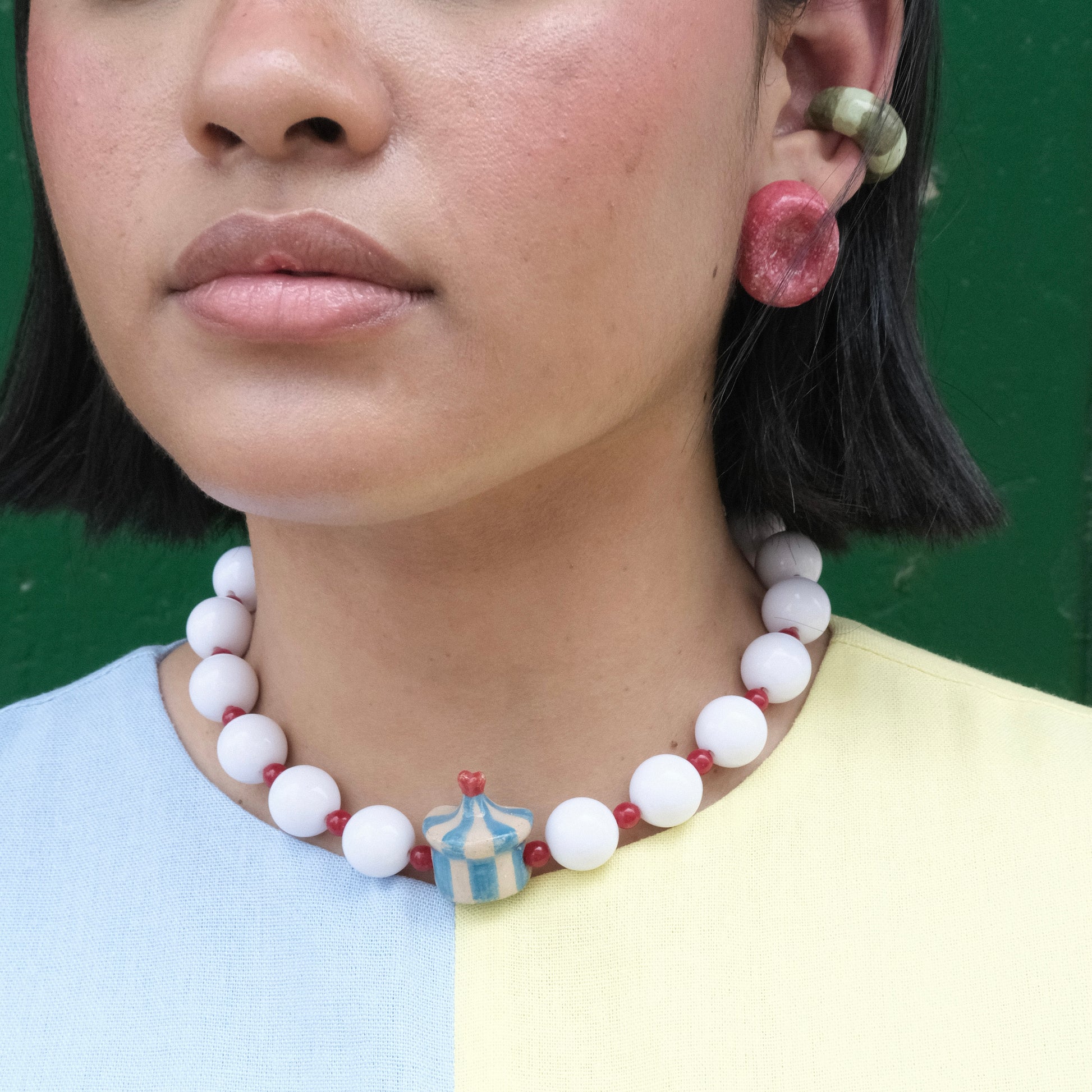 Rostro y cuello de modelo pluszize de tes morena y cabello corto color negro, usando vestido amarillo y celeste, collar Dulcero Festival estilo chunky, aretes deliciosos y earcuff Cidra en colores verde claro, verde oscuro, rojo, azul y blanco en material de cerámica. 