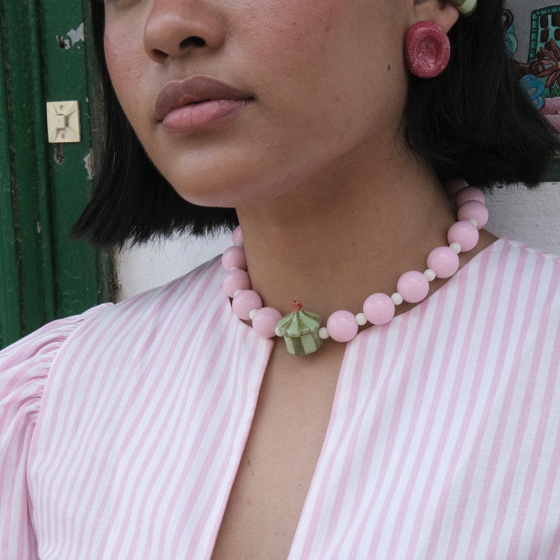 Rostro y cuello de modelo pluszize de tes morena   cabello corto color negro, usando top de rayas en colores blanco y rosado, Collar Dulcero Cidra estilo chunky y aretes deliciosos en colores rojo, verde claro y verde oscuro en material de cerámica. 