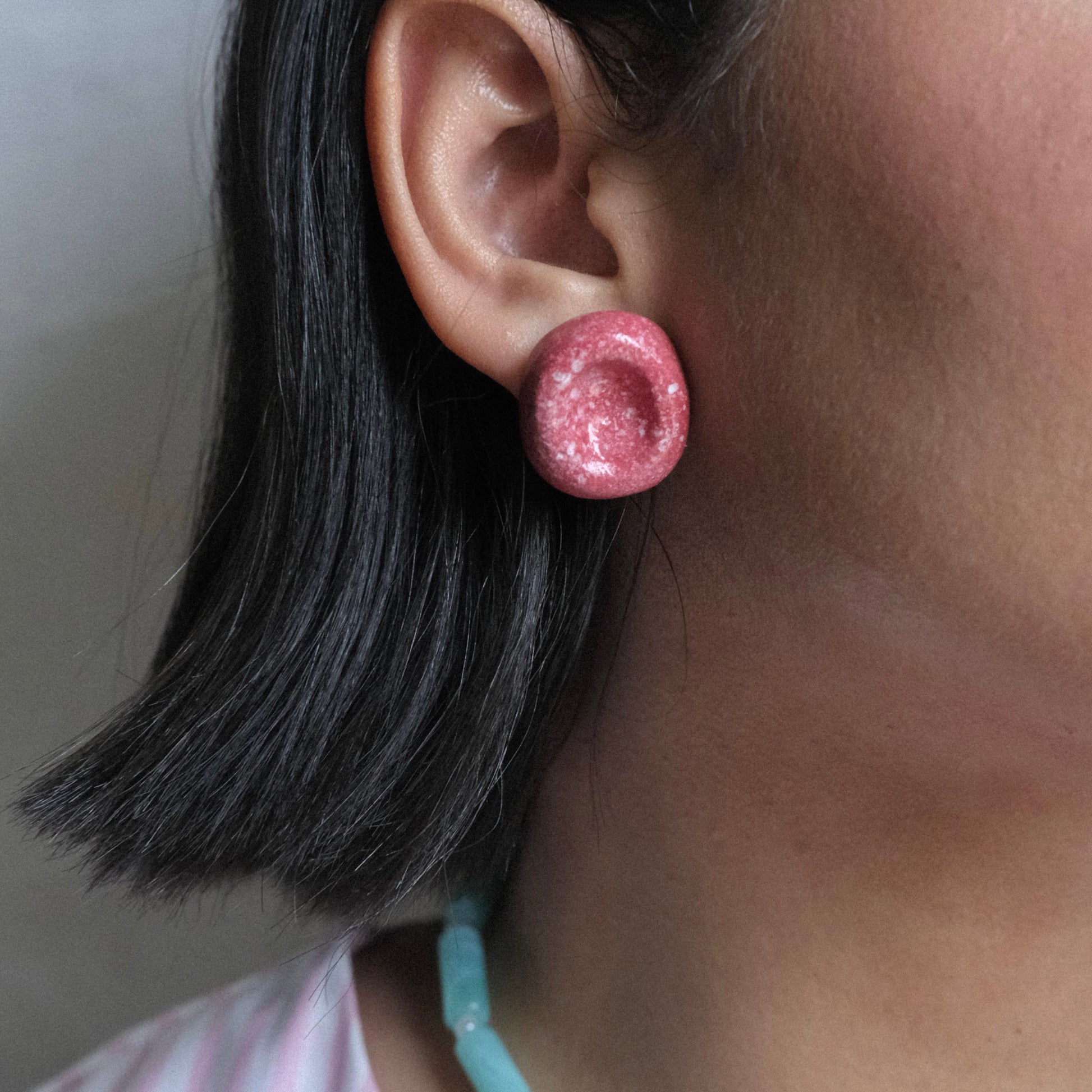 Oreja de modelo de tes morena y cabello corto y negro en fondo blanco, usando aretes Delicioso Bocadillo estilo topo en material de cerámica color rojo.