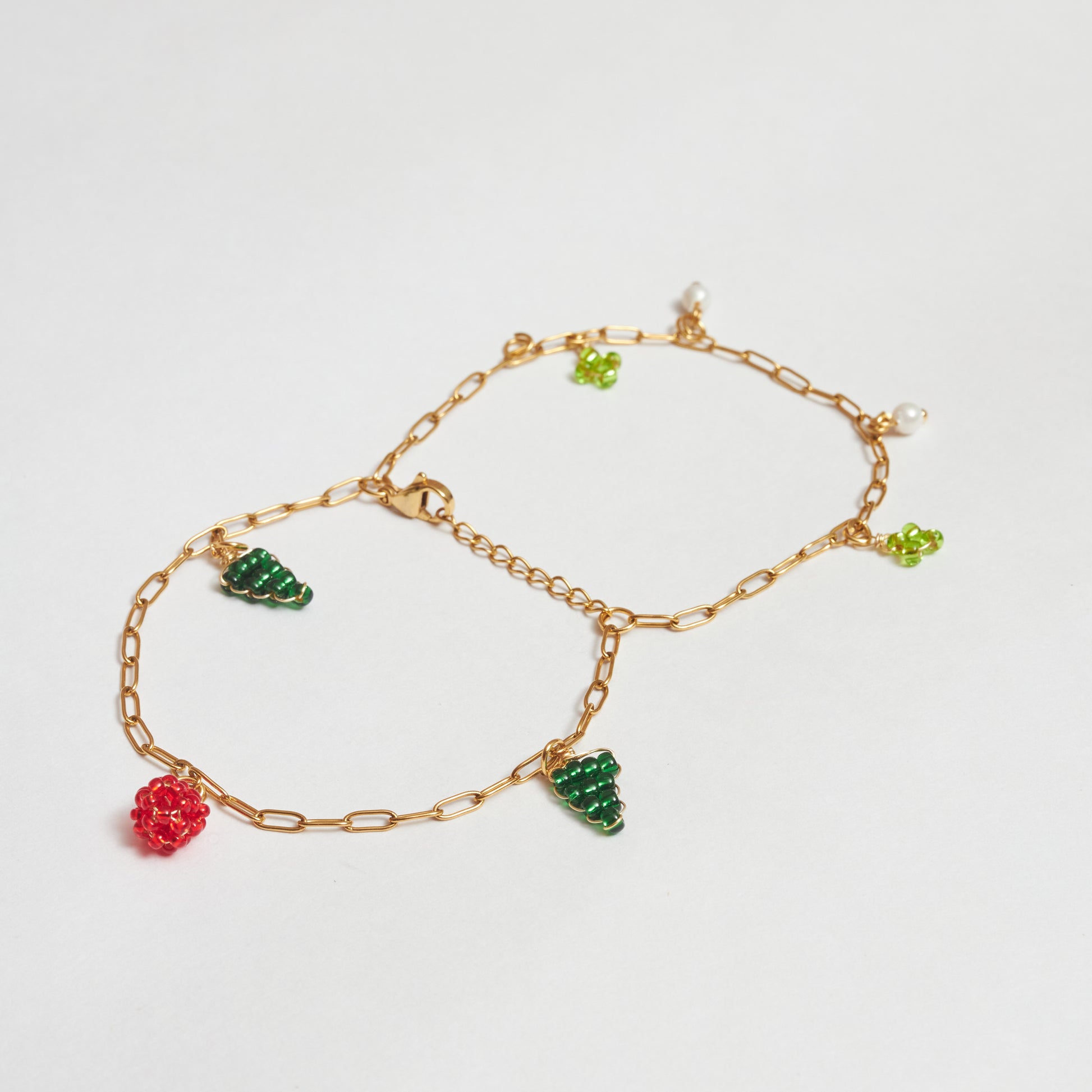 brazalete de doble cadena abierto en estilo mariposa, en material de acero inoxidable color dorado con dijes de perlas blancas y en mostacilla tejida  con forma de árbol, flor y fruto rojo en colores verde claro, verde oscuro y rojo. 
